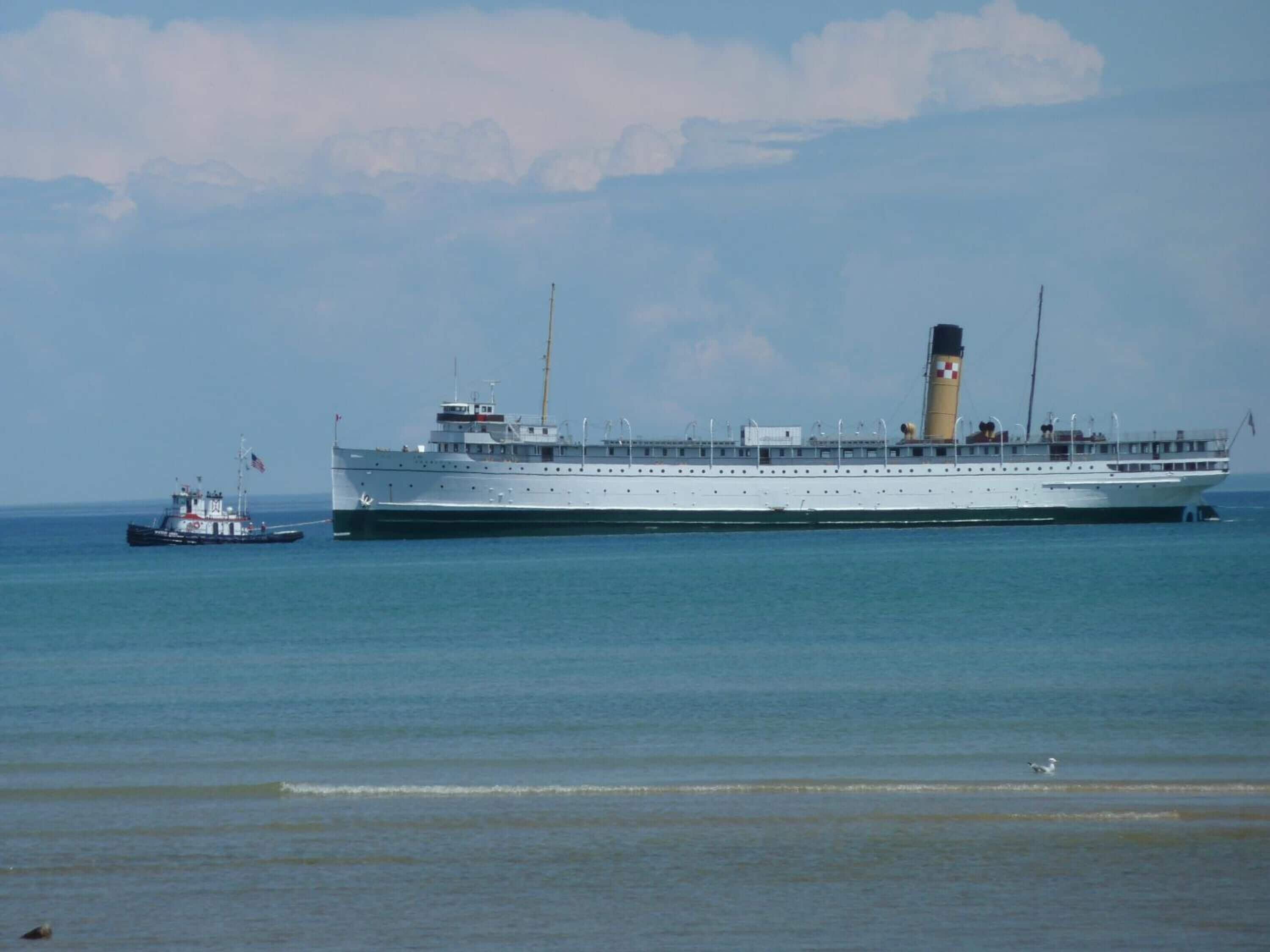 A large cruise ship with a prominent funnel and multiple decks, docked at a port with a smaller tugboat nearby.