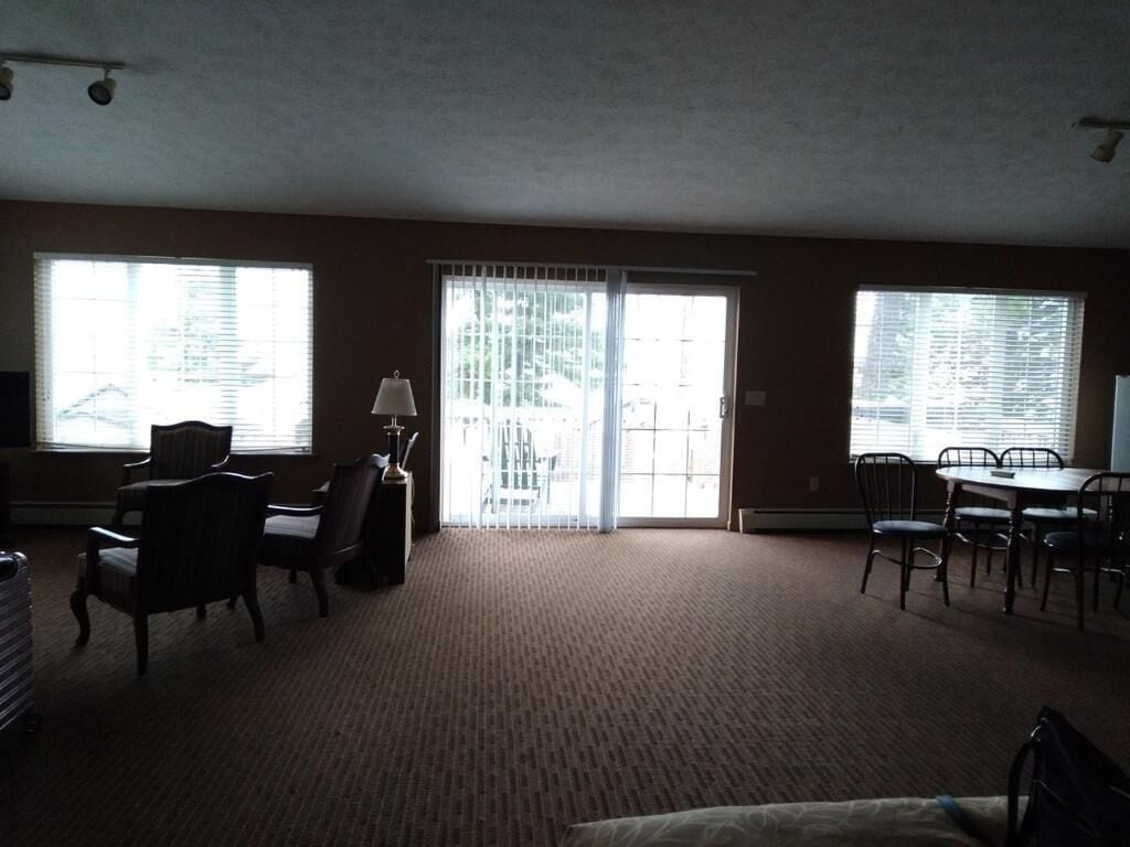 A spacious living room with a carpeted floor, a dining table with chairs, and large windows with blinds.