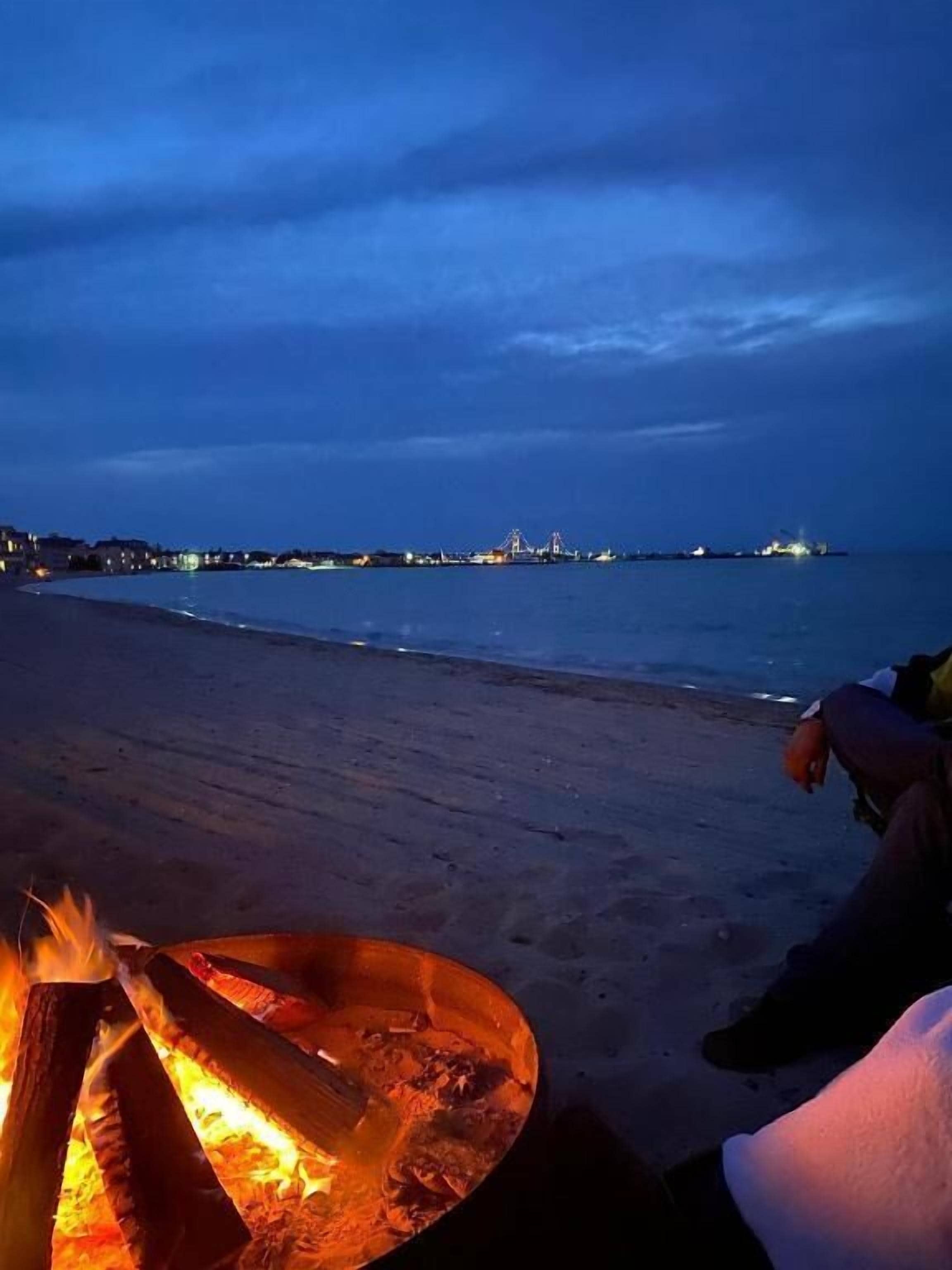 A beach scene with a fire pit and a person sitting by the water.