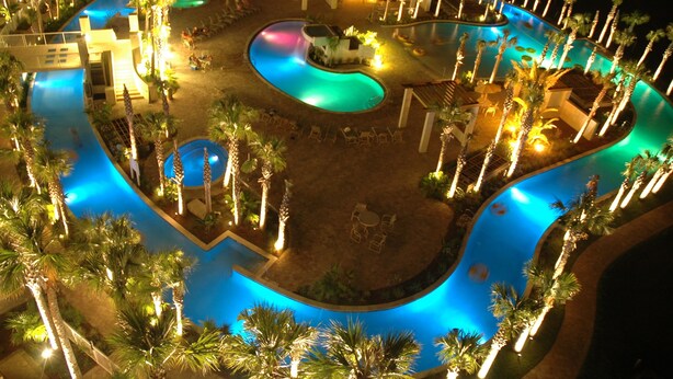 A nighttime view of a resort pool area with multiple swimming pools, palm trees, and lounge chairs. - Destin West Vacations (Fort Walton Beach)