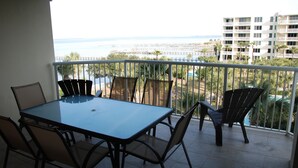A balcony with a glass table and chairs, overlooking a beach and a resort area with palm trees and a pool. - Destin West Vacations (Fort Walton Beach)