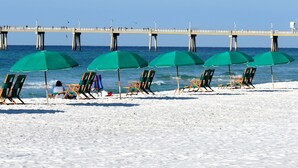 A beach with rows of green umbrellas and beach chairs, a pier in the background, and clear blue skies. - Destin West Vacations (Fort Walton Beach)