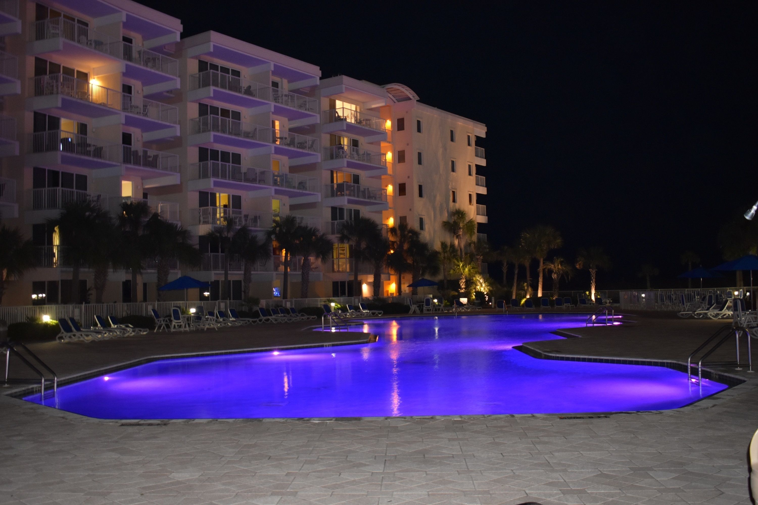 Et hotel med flere balkoner, en swimmingpool med blå belysning og palmer.