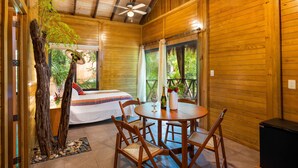 Interior de una cabaña de madera con una cama, una mesa de comedor, sillas, ventiladores de techo y vistas a la vegetación.