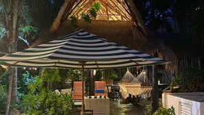 Una estructura con techo de paja, con una mesa y sillas bajo un paraguas a rayas, rodeada de plantas tropicales.