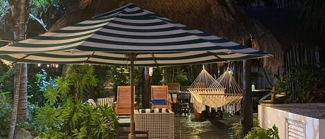 A thatched-roof structure with a table and chairs under a striped umbrella, surrounded by tropical plants.