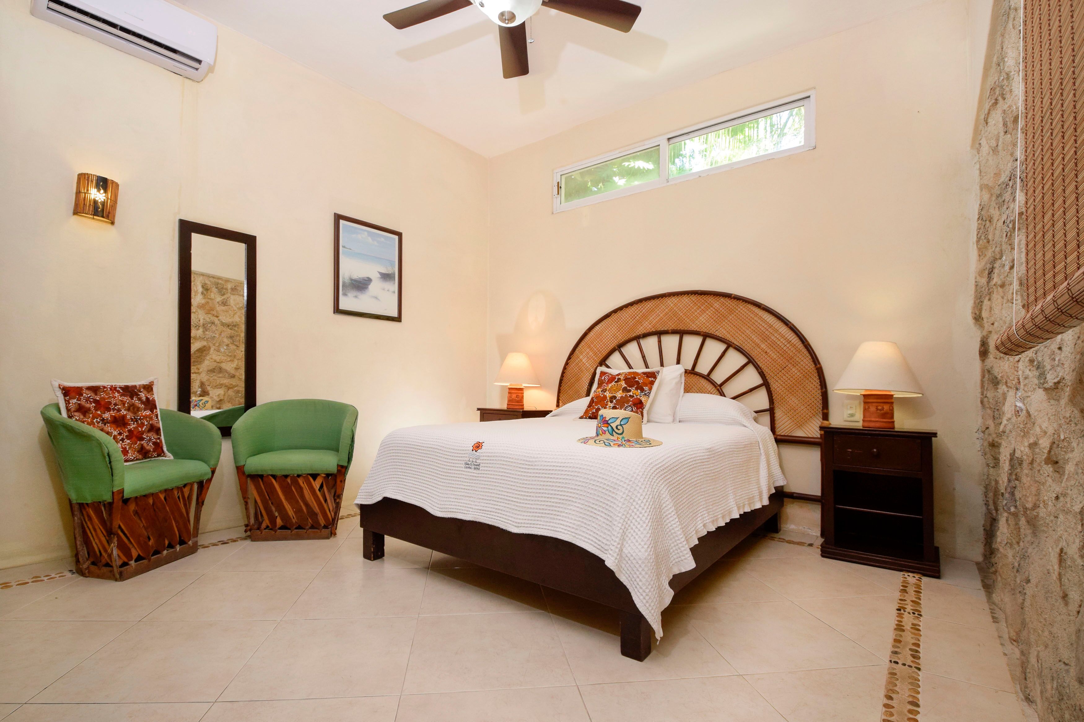 A bedroom with a bed, two green chairs, a wooden headboard, two bedside tables with lamps, and a ceiling fan.