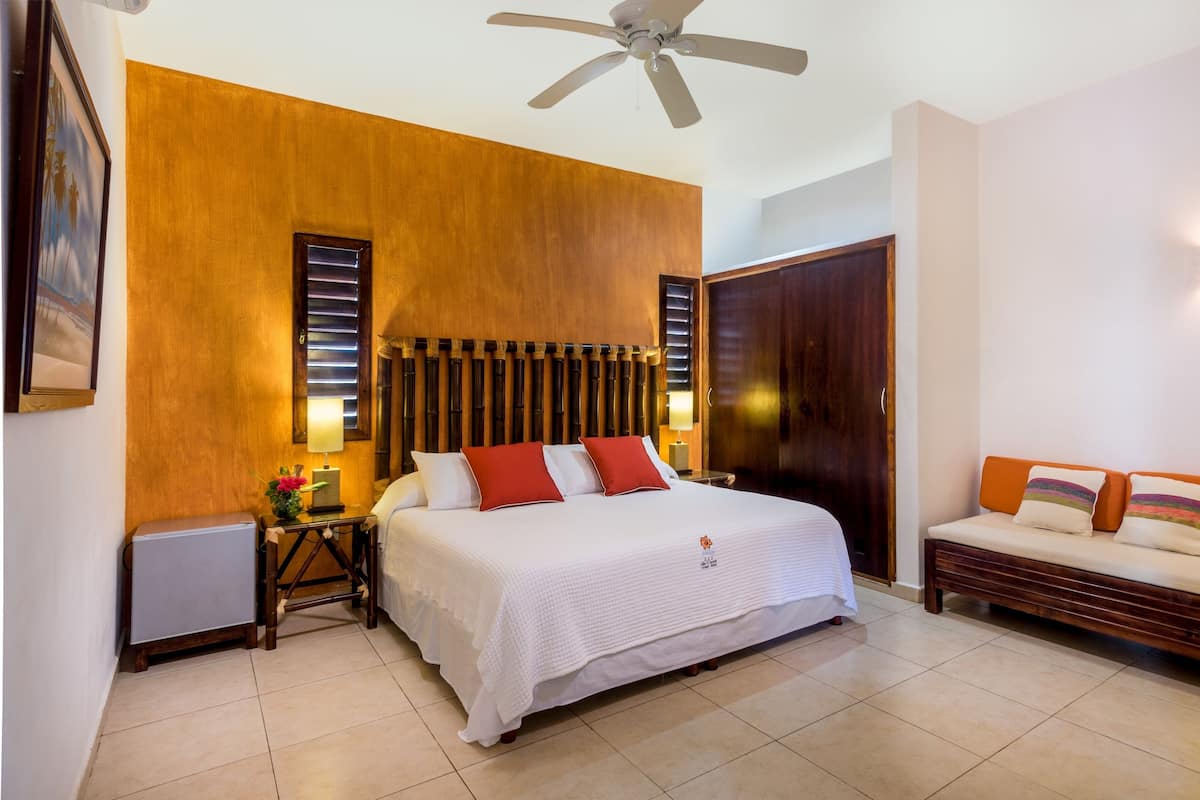 A hotel room with a bed, a nightstand, a lamp, a ceiling fan, a wooden wall, a door, and a sofa.