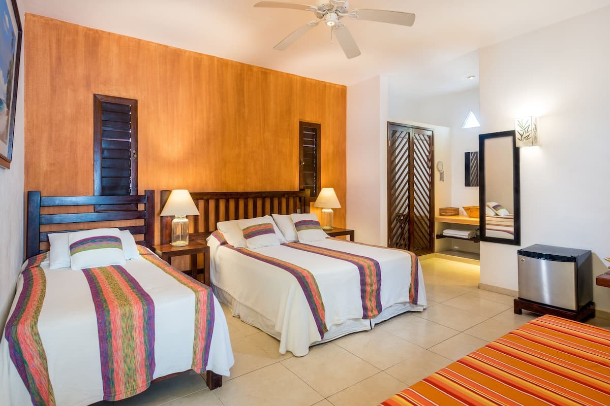 A hotel room with two beds, wooden headboards, a ceiling fan, a bathroom with a shower, and a small table with a lamp.