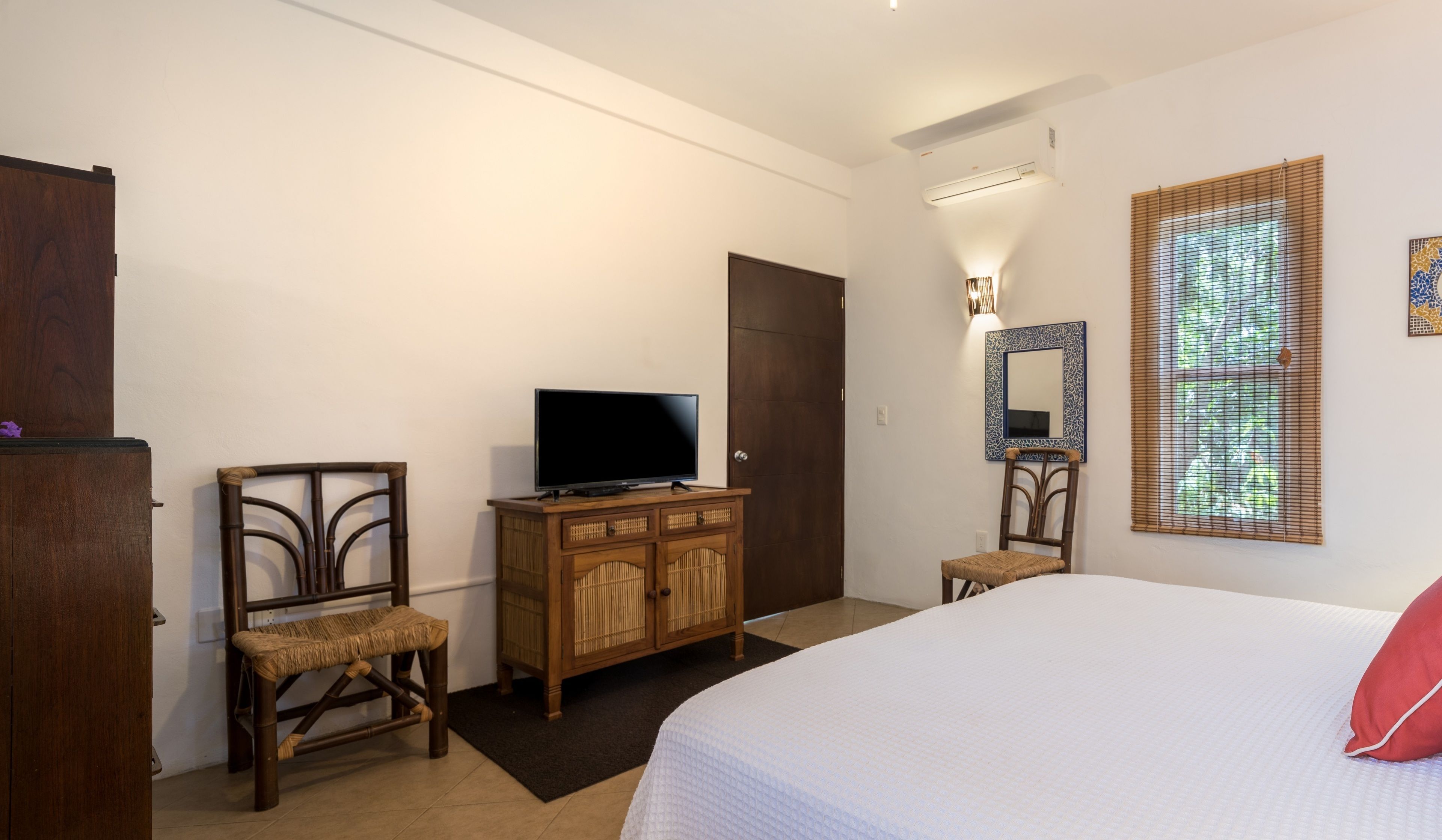 Une chambre à coucher avec un lit, un téléviseur posé sur une commode en bois, une chaise, une petite table et une porte.