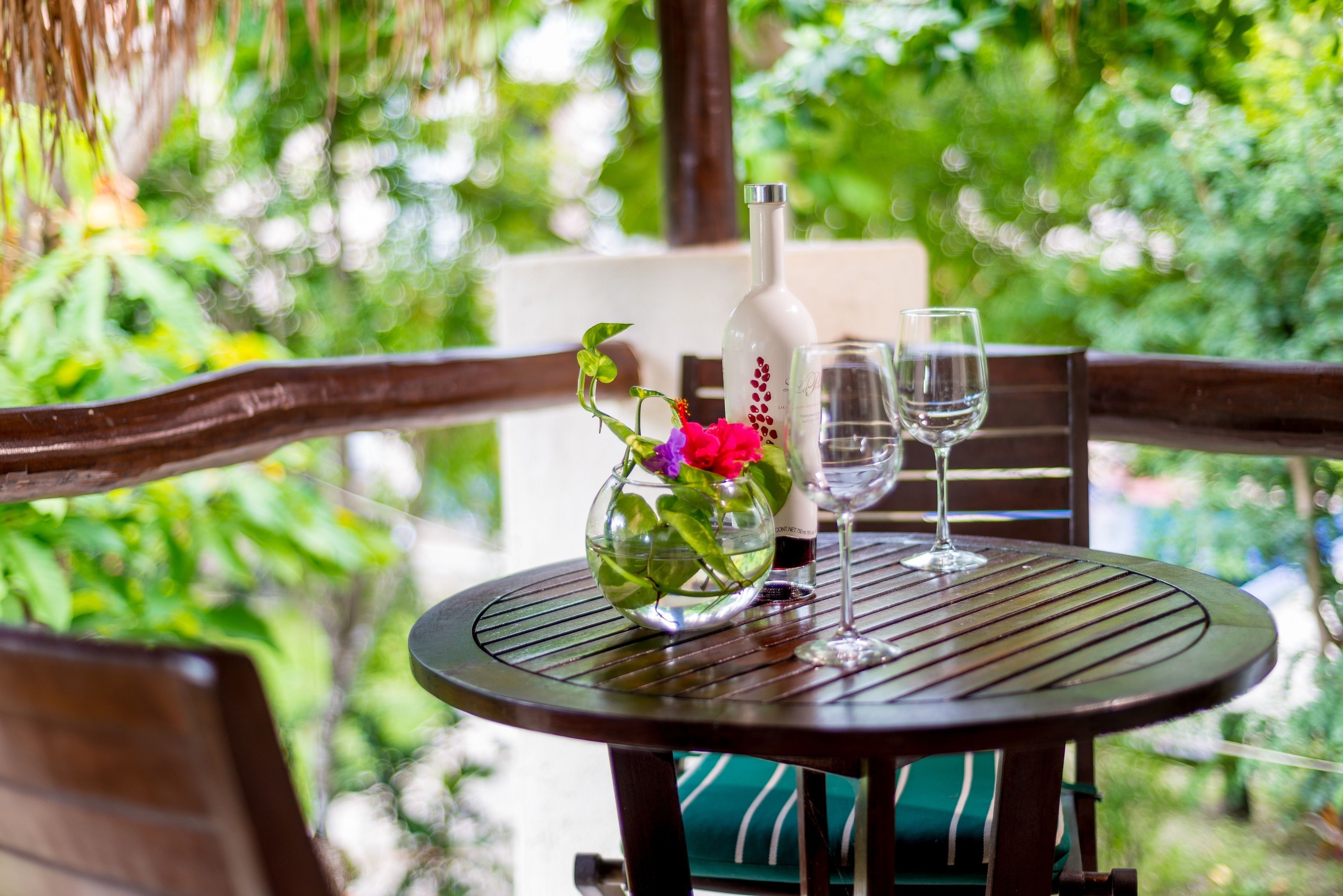 A round table with a bottle and two glasses, surrounded by wooden chairs with green cushions.