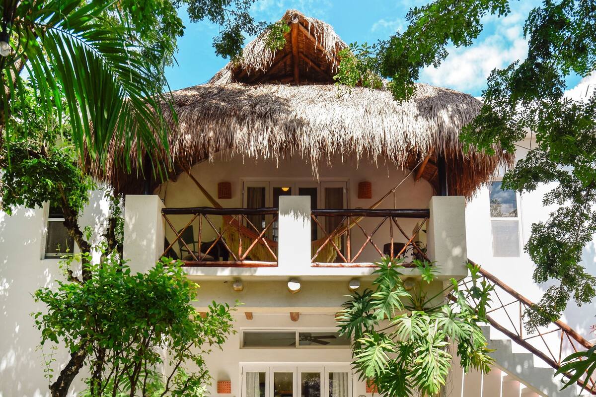 A two-story building with a thatched roof, surrounded by lush greenery and a gravel pathway leading to the entrance.