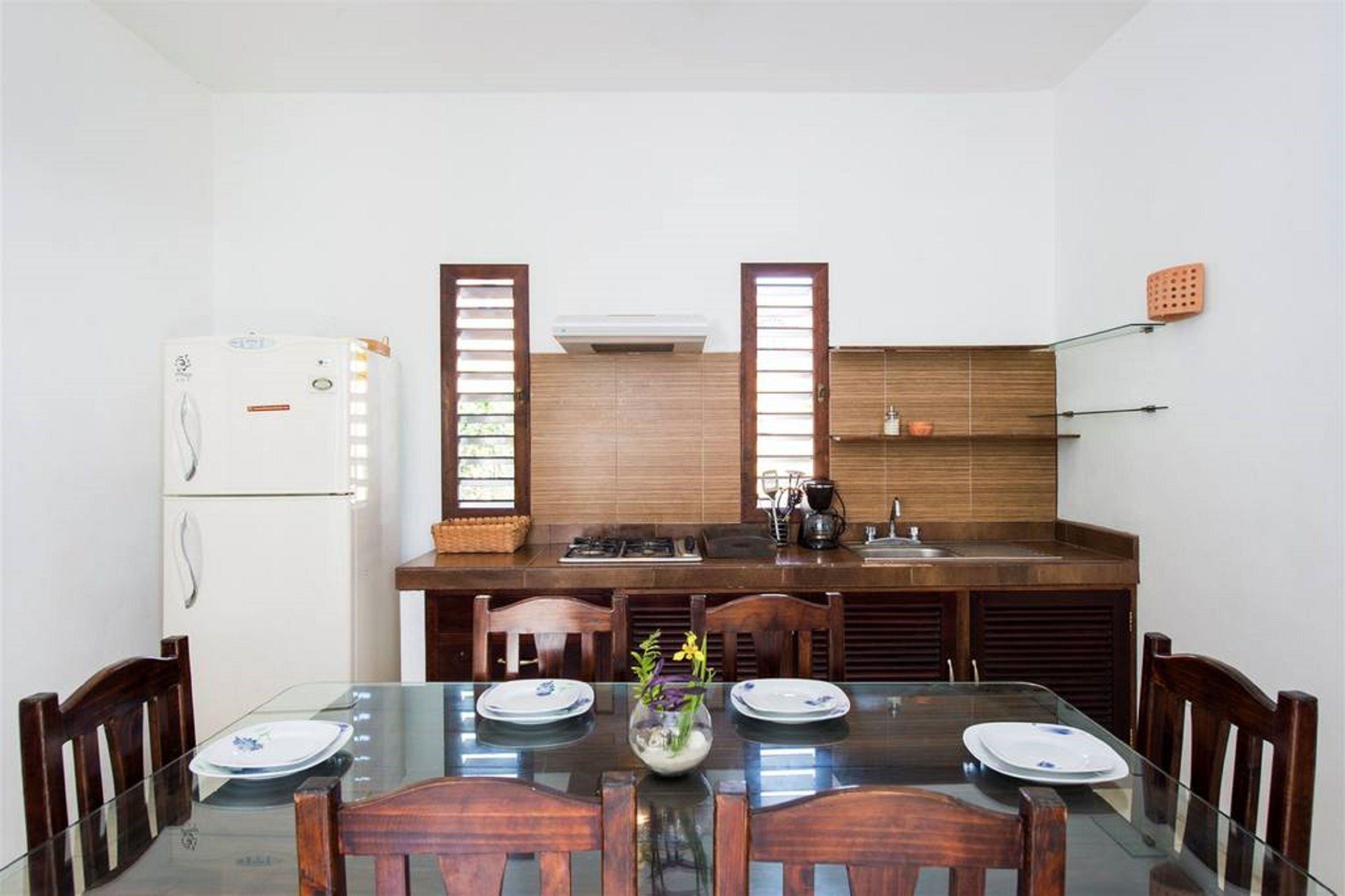 A dining area with a glass table, wooden chairs, and a kitchen in the background with a refrigerator, stove, and cabinets.