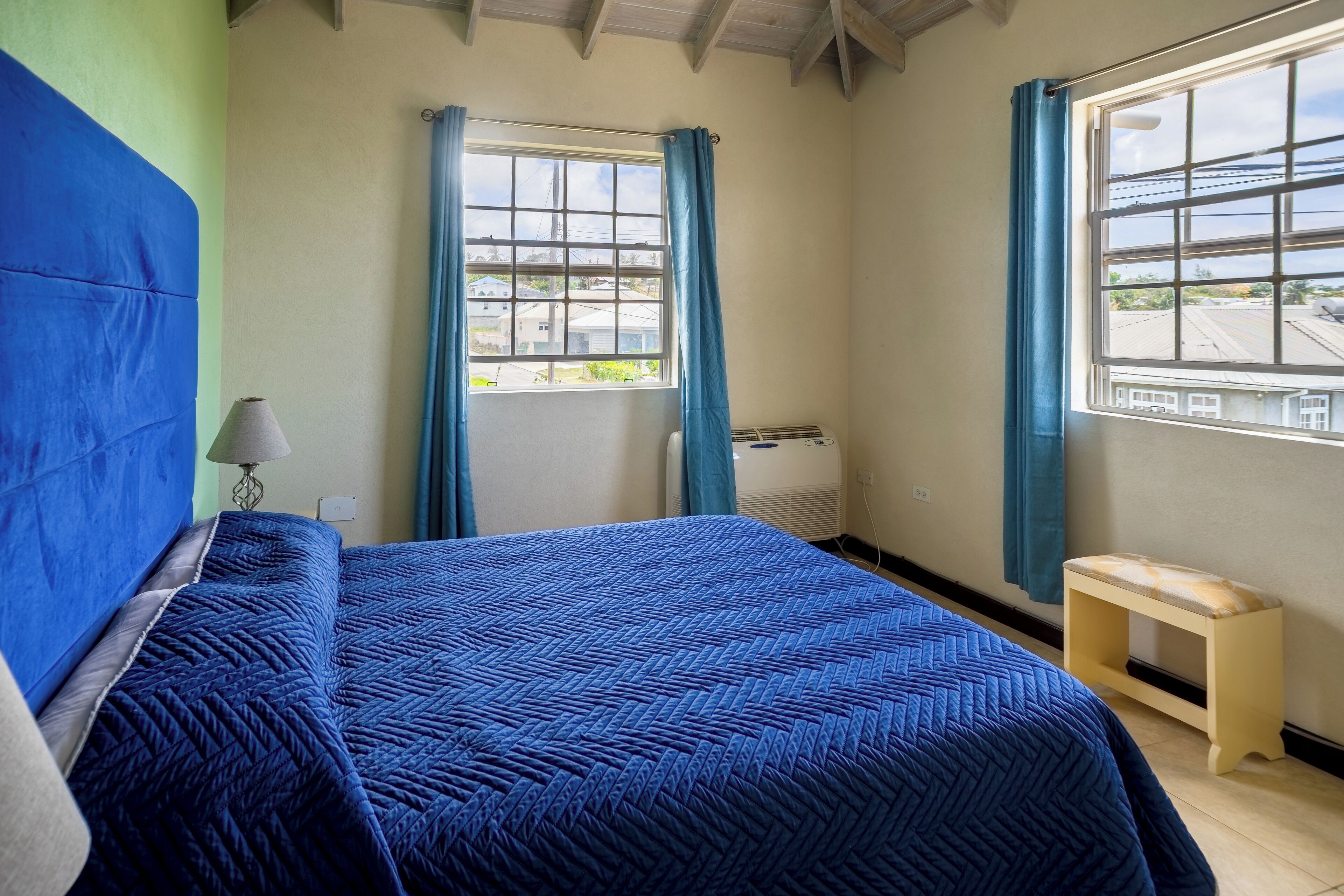 Un dormitorio con una cama grande, dos ventanas con cortinas, una mesita de noche y una lámpara.