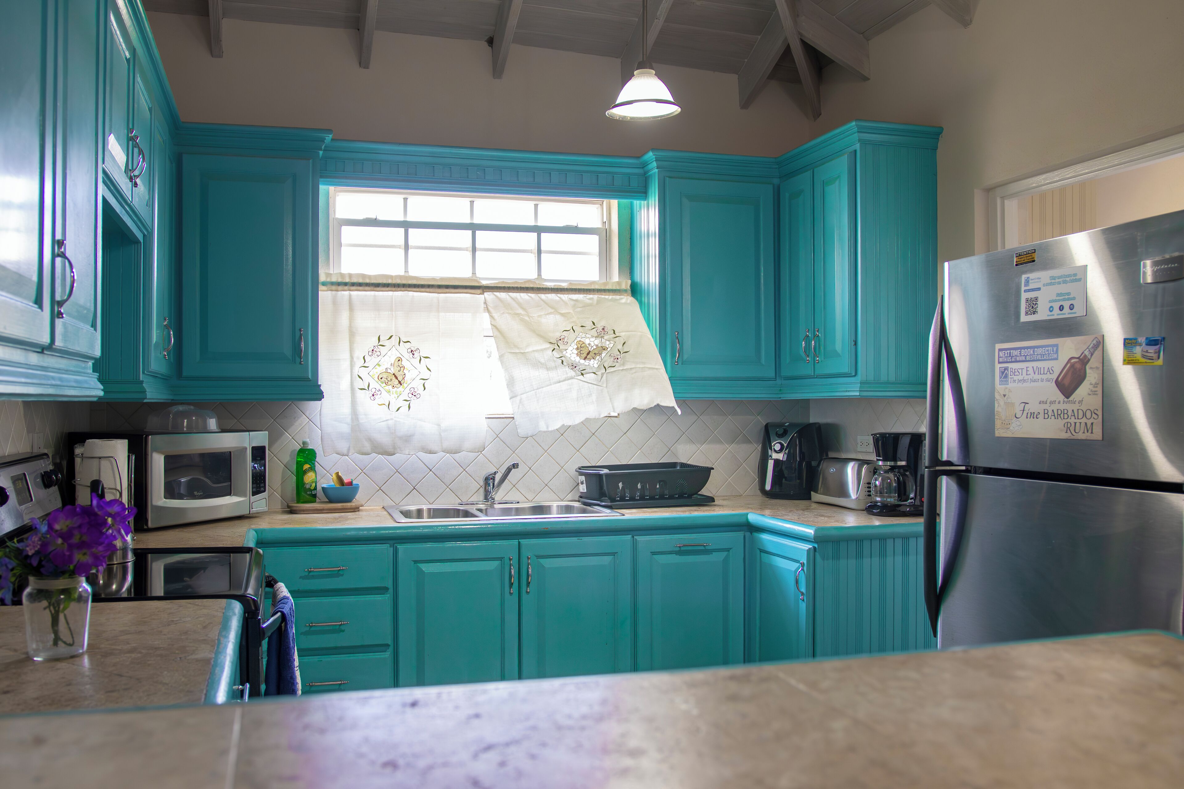 A kitchen with teal cabinets, a stainless steel refrigerator, a microwave, and a window with a floral curtain.
