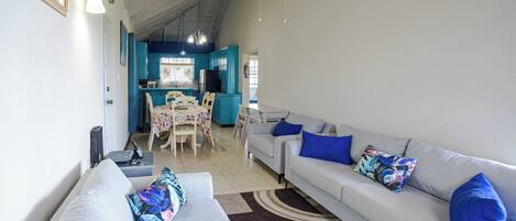 A spacious living area with a sofa set, a dining table with chairs, and a kitchen in the background.
