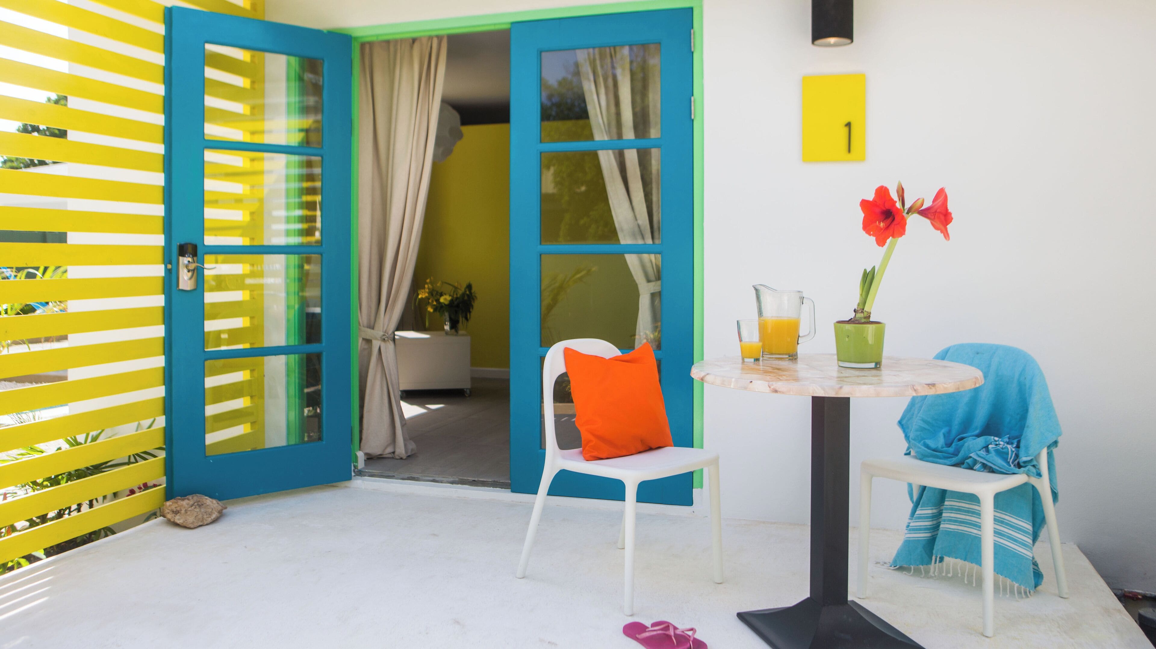 A bright room with a blue door, a white chair, a small table with a pitcher of orange juice, and a vase with red flowers.