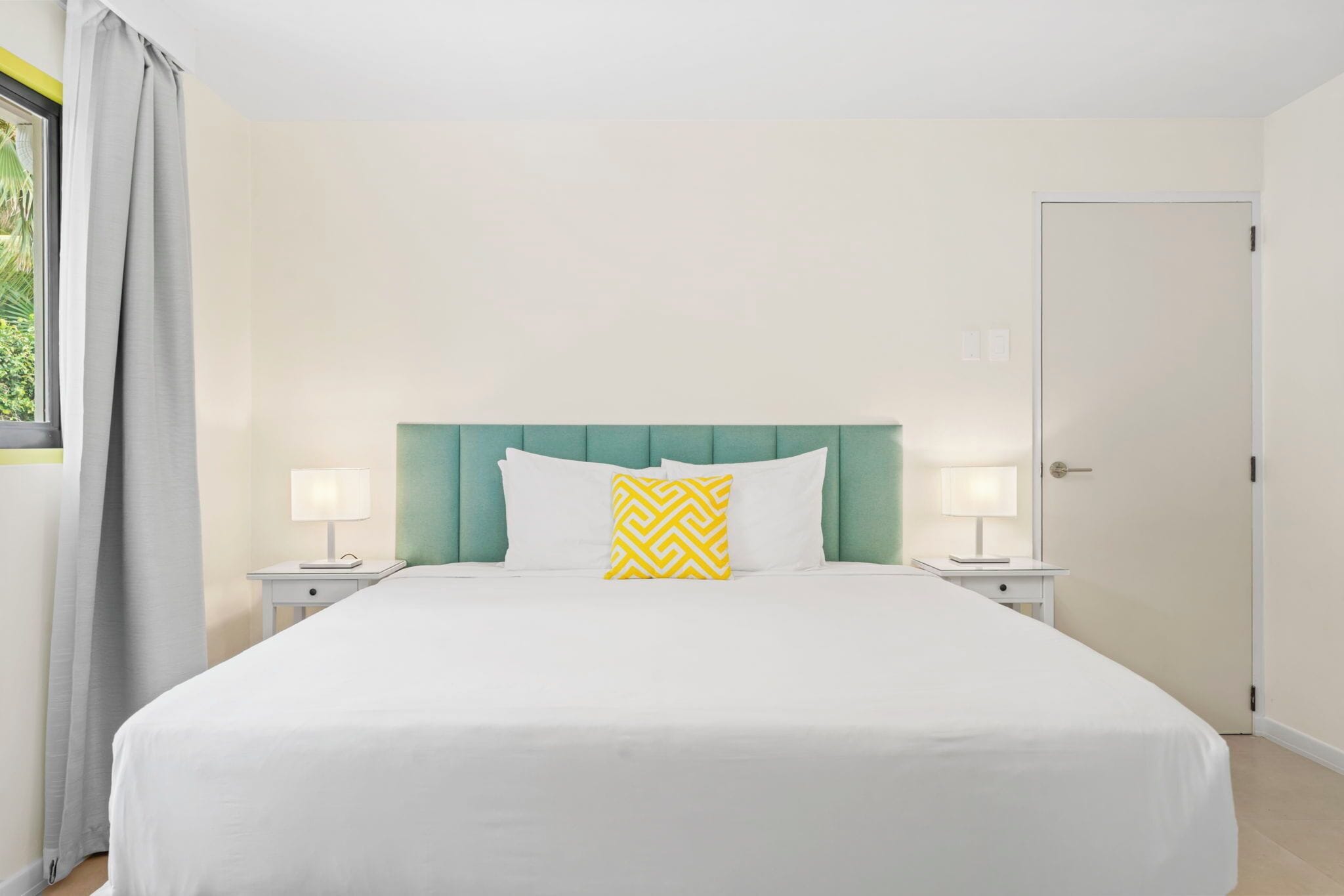 Una cama bien hecha con sábanas blancas y cabecera verde, dos lámparas de noche y una puerta a la derecha.