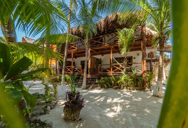 A traditional wooden structure with a thatched roof, surrounded by palm trees and tropical plants.