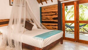A bedroom with a bed, a mosquito net, a wooden headboard, a wooden door with a view, and a hammock outside.