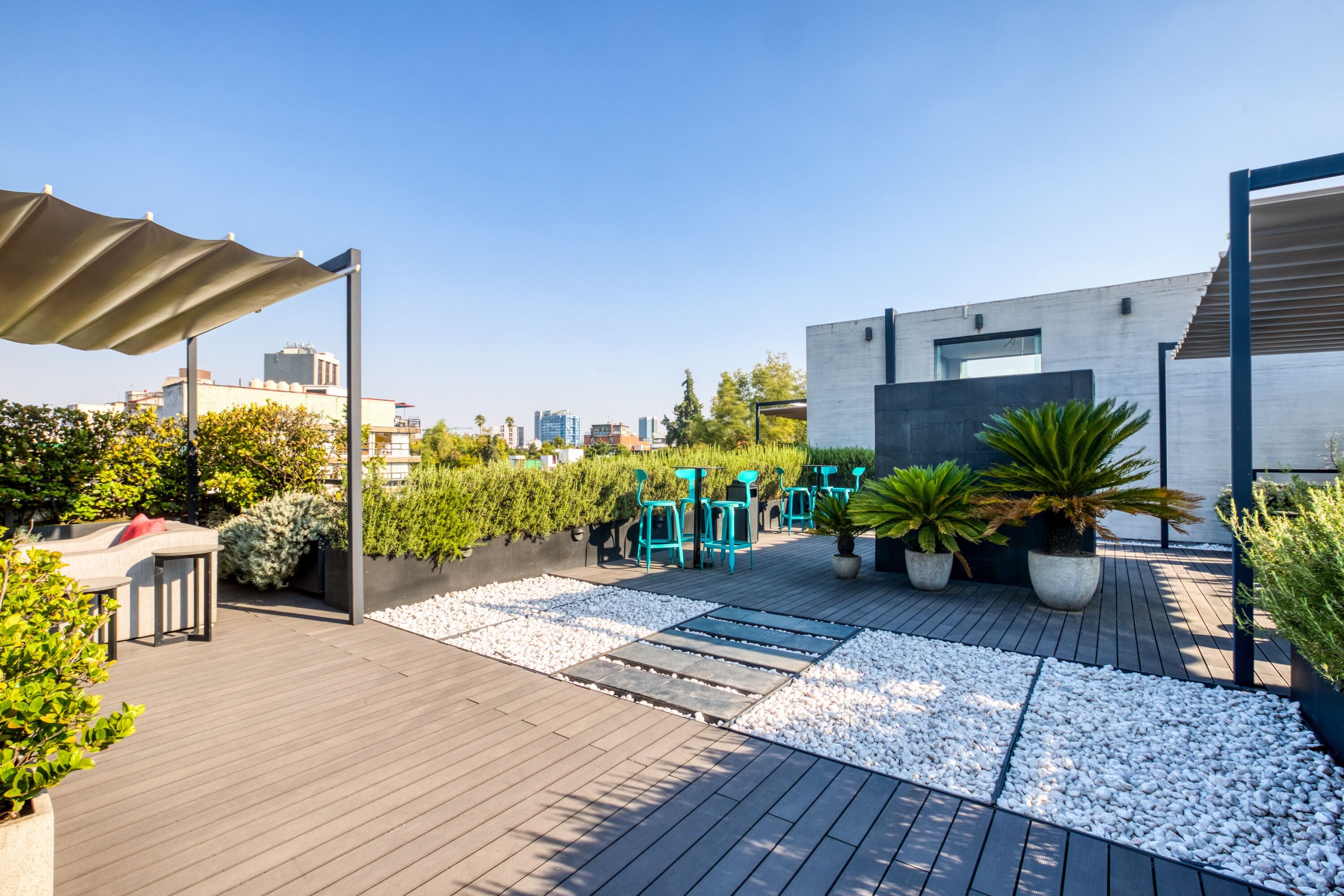 A rooftop patio with a wooden deck, seating area, potted plants, and a modern building in the background.