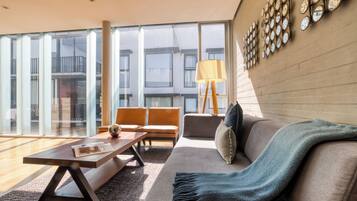 A modern living room with a sofa, armchairs, a coffee table, and a lamp. Large windows offer a view of the outside.