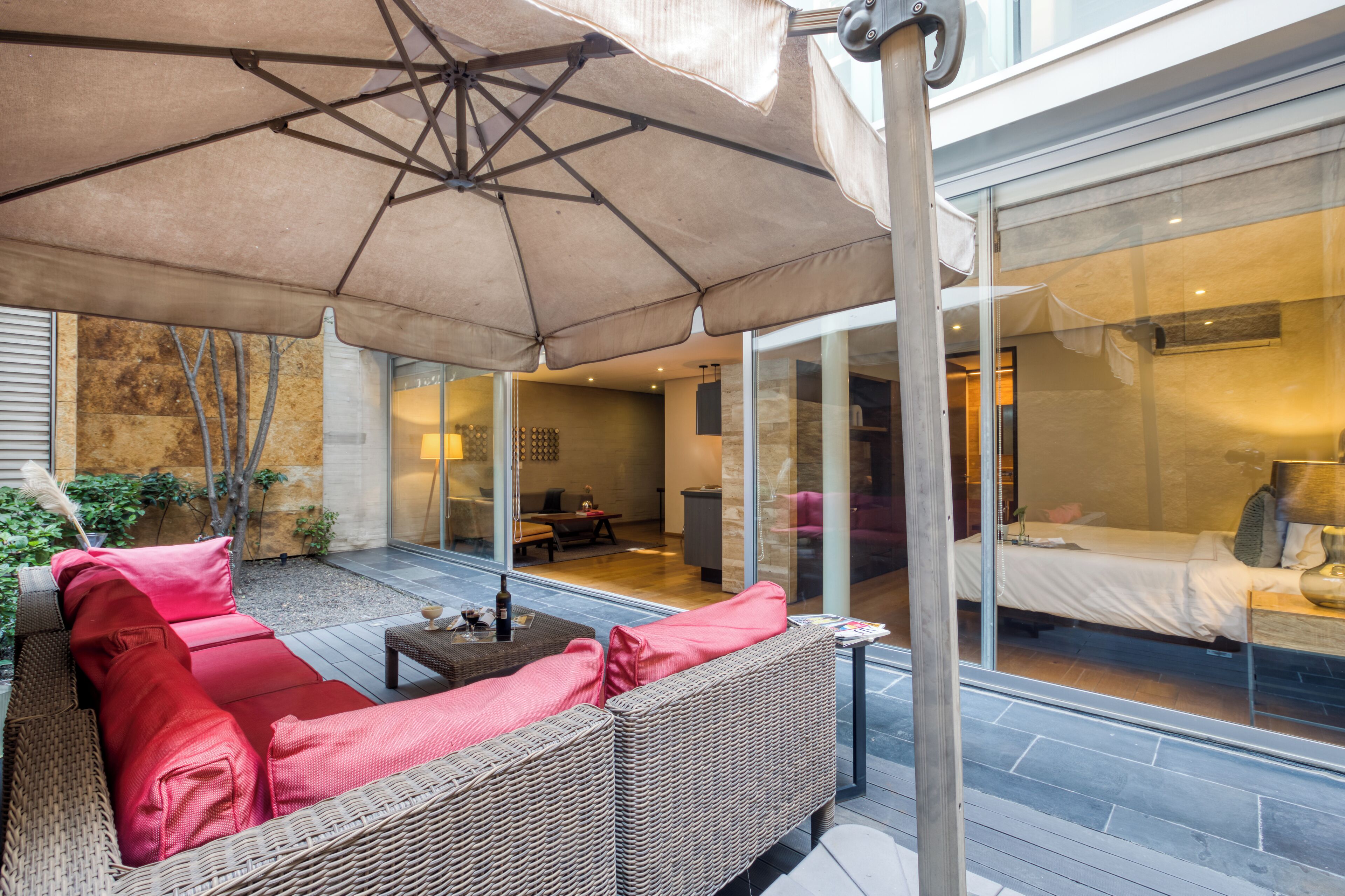 A modern outdoor patio with wicker furniture, a large umbrella, and a glass-enclosed bedroom area.