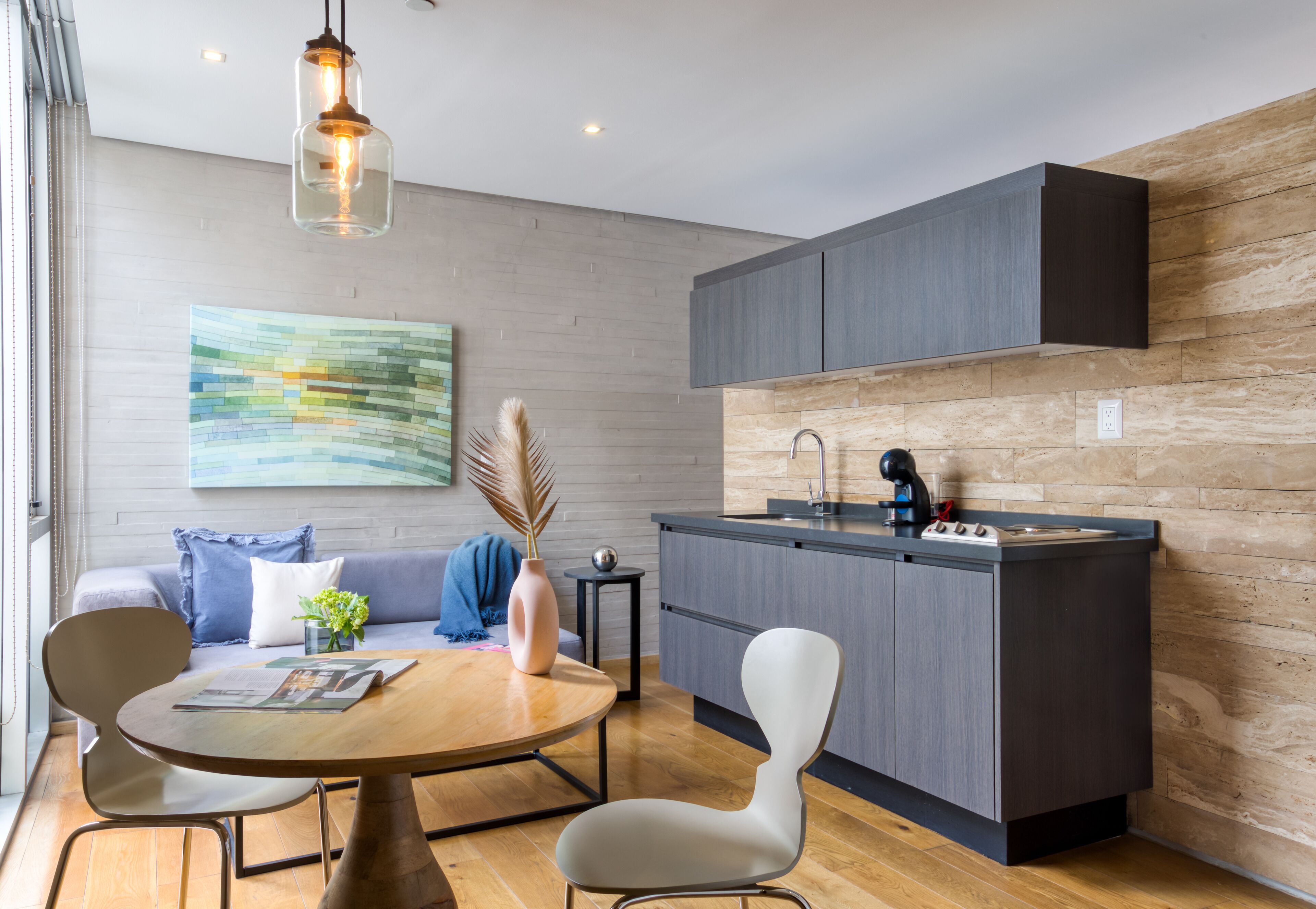 Una cocina moderna con armarios grises, encimera de madera y estufa integrada. Hay una zona de comedor con una mesa redonda y sillas, un jarrón con flores y un cuadro en la pared.