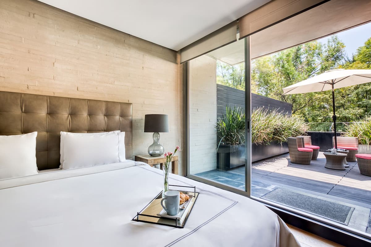 A modern hotel room with a large bed, a bedside table with a lamp, and a view of the outdoor patio with seating and plants.