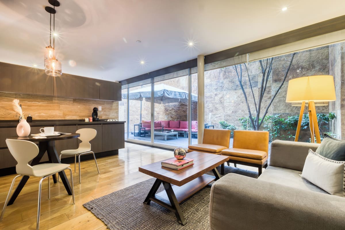 A modern living room with a sofa, chairs, a coffee table, and a lamp. There is a kitchen area with a dining table and chairs. Large windows provide a view of the outdoors.