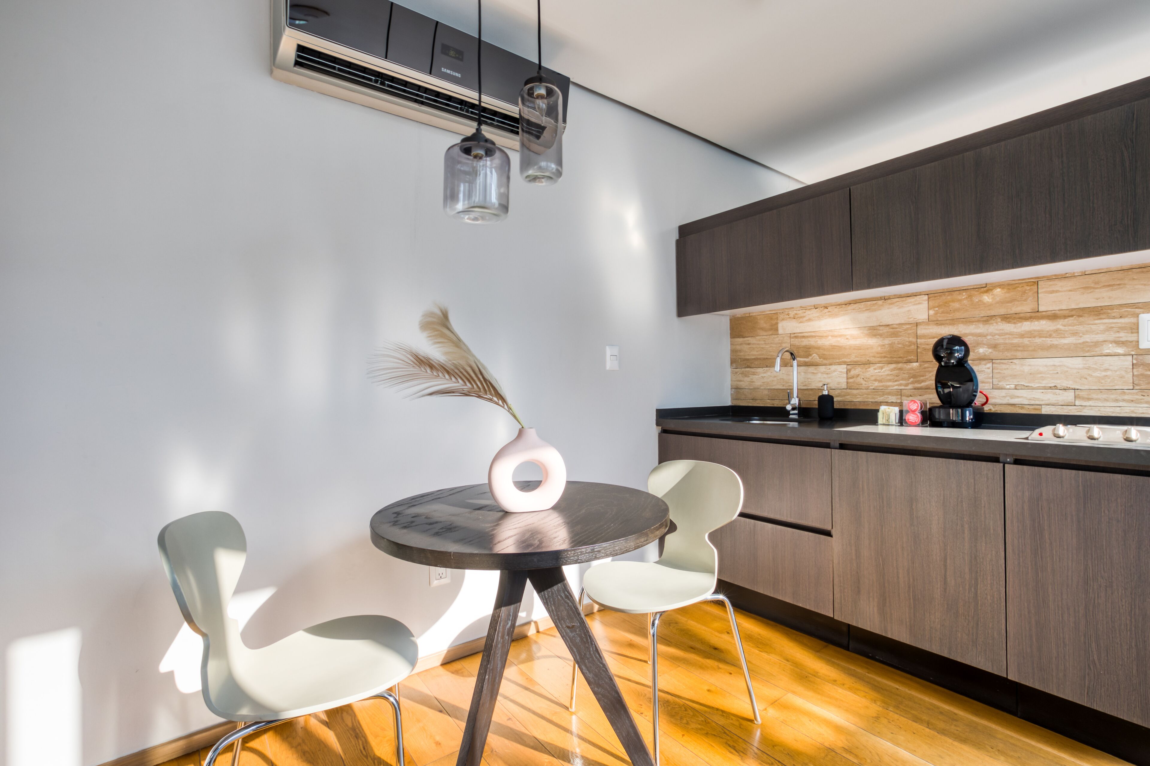 Une cuisine moderne avec des meubles de rangement en bois foncé, une table de salle à manger ronde accompagnée de deux chaises et des luminaires suspendus.