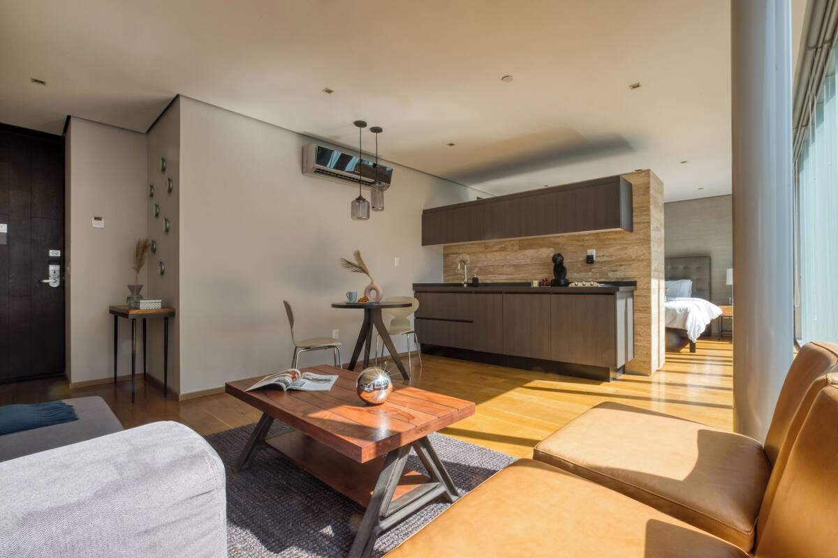 A modern living room with a wooden coffee table, a sofa, and a leather armchair. A kitchen with wooden cabinets and a built-in oven is visible in the background.