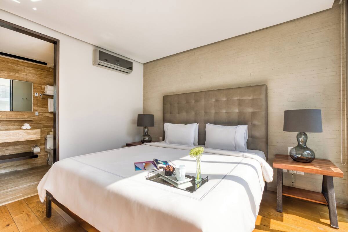 A hotel room with a large bed, two bedside tables with lamps, a wooden headboard, a wooden vanity with a mirror, and a small window.