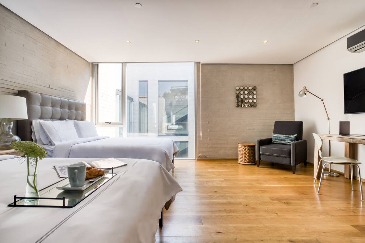 A modern hotel room with two beds, a desk, a chair, and a large window offering a city view.