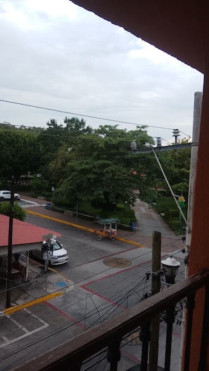A view from a balcony overlooking a street with a parked car and a motorcycle, trees, and a building with a red roof.
