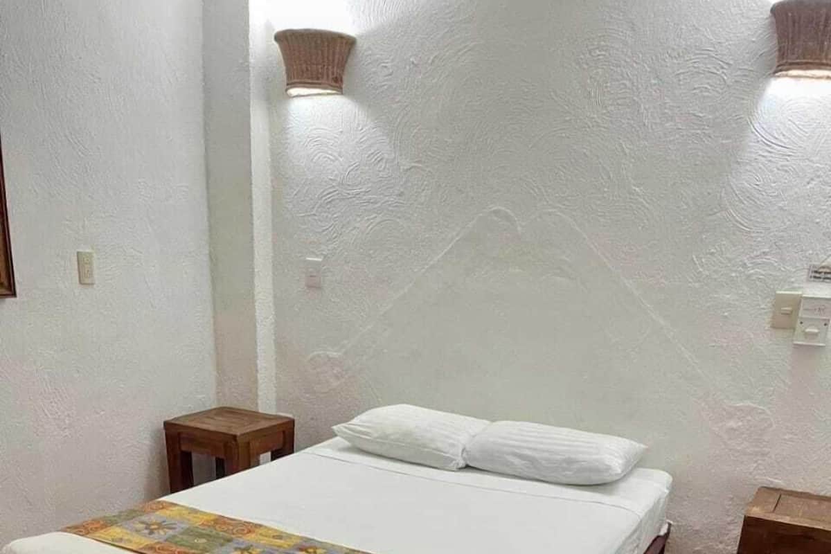 Une chambre avec un lit simple, une tête de lit en bois, une couvre-lit à motifs, une petite table de chevet en bois et un luminaire fixé au mur.