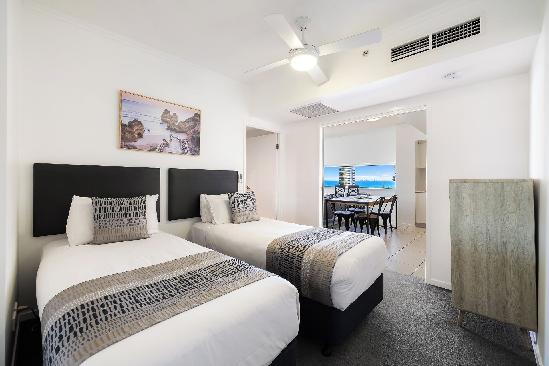 Una camera d'albergo con due letti, una ventola a soffitto, un quadro e vista mare.