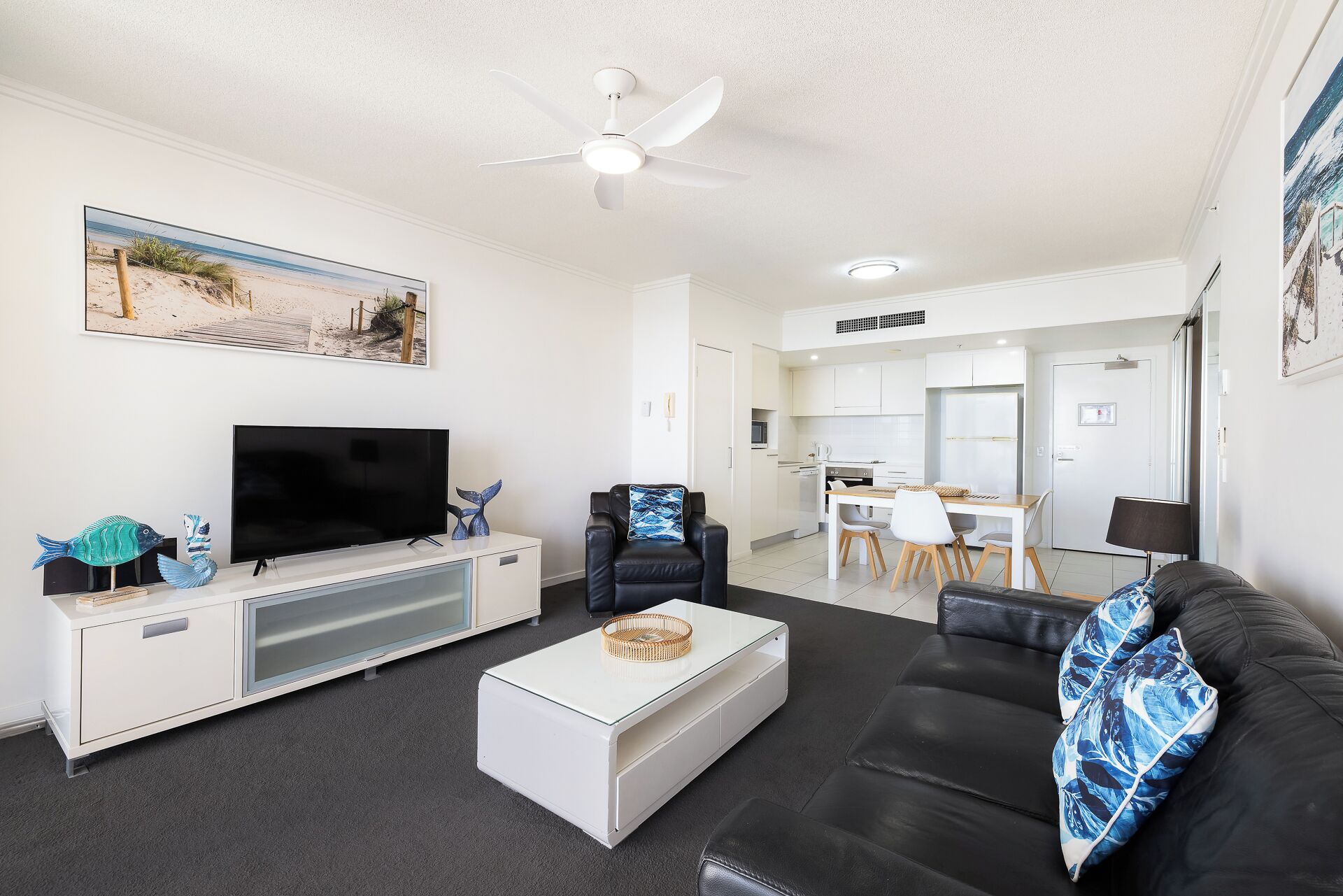Ein modernes Wohnzimmer mit Flachbildfernseher, einer schwarzen Ledersofa und einem Couchtisch. Es gibt einen Deckenventilator, eine Küchenzeile im Hintergrund und ein Strandbild an der Wand.