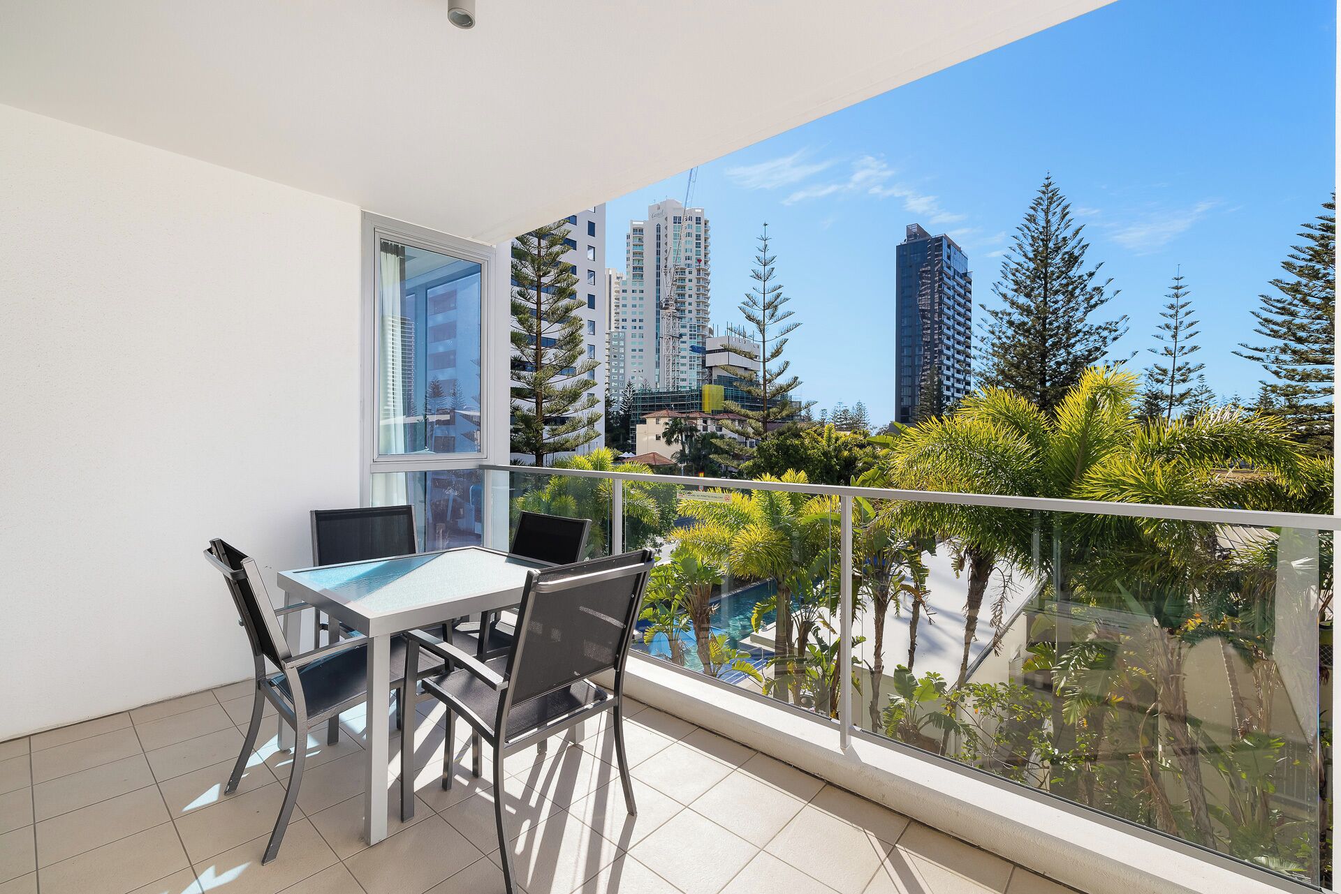 A balcony with a table and chairs, a window, and a view of tall buildings and trees.