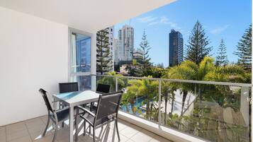 A balcony with a table and chairs, a window, and a view of tall buildings and trees.