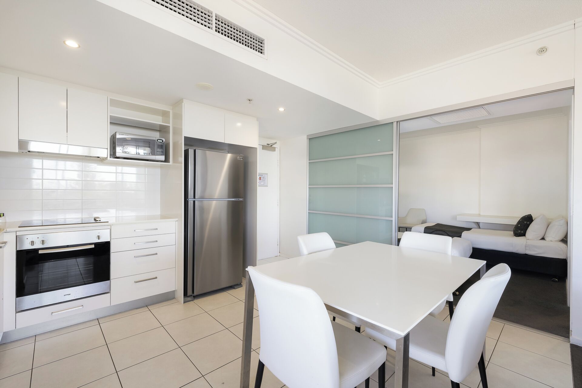 A modern kitchen with white cabinets, a stainless steel refrigerator, a microwave, and an oven. There is a dining area with a white table and chairs.