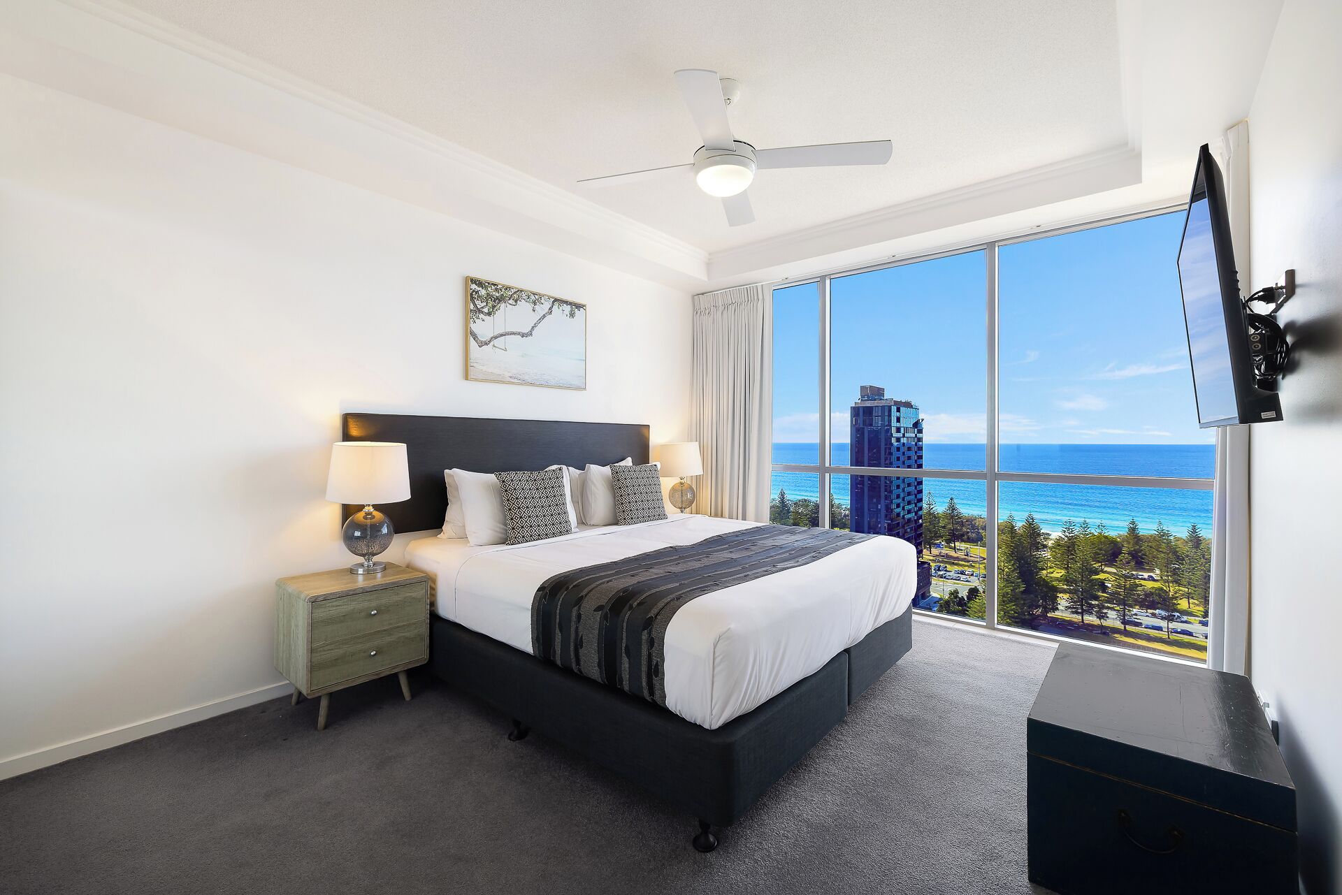 A modern hotel room with a large bed, a ceiling fan, a TV, and a view of the ocean and city skyline.