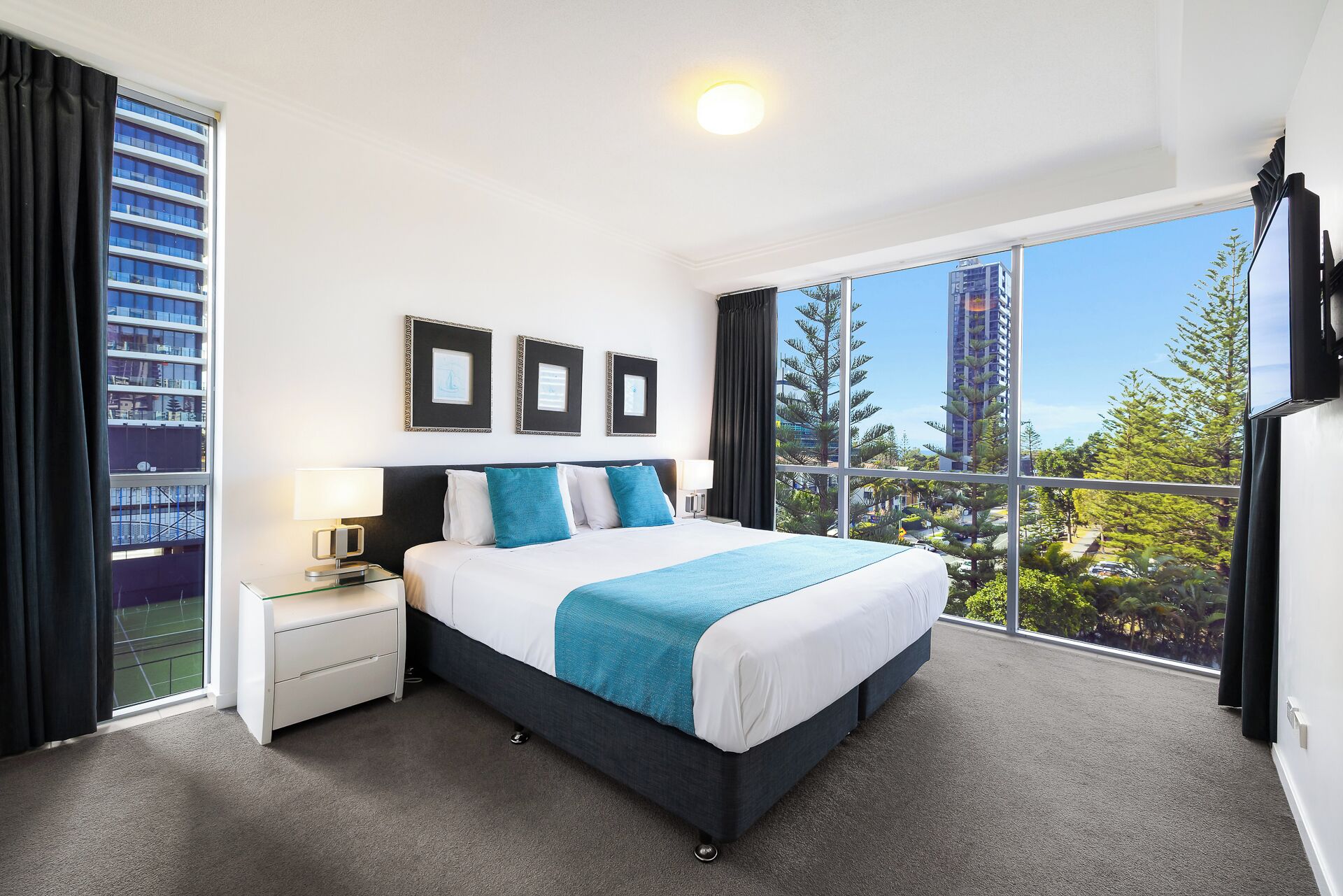 A modern hotel room with a large bed, a bedside table, a TV, and a view of the cityscape.