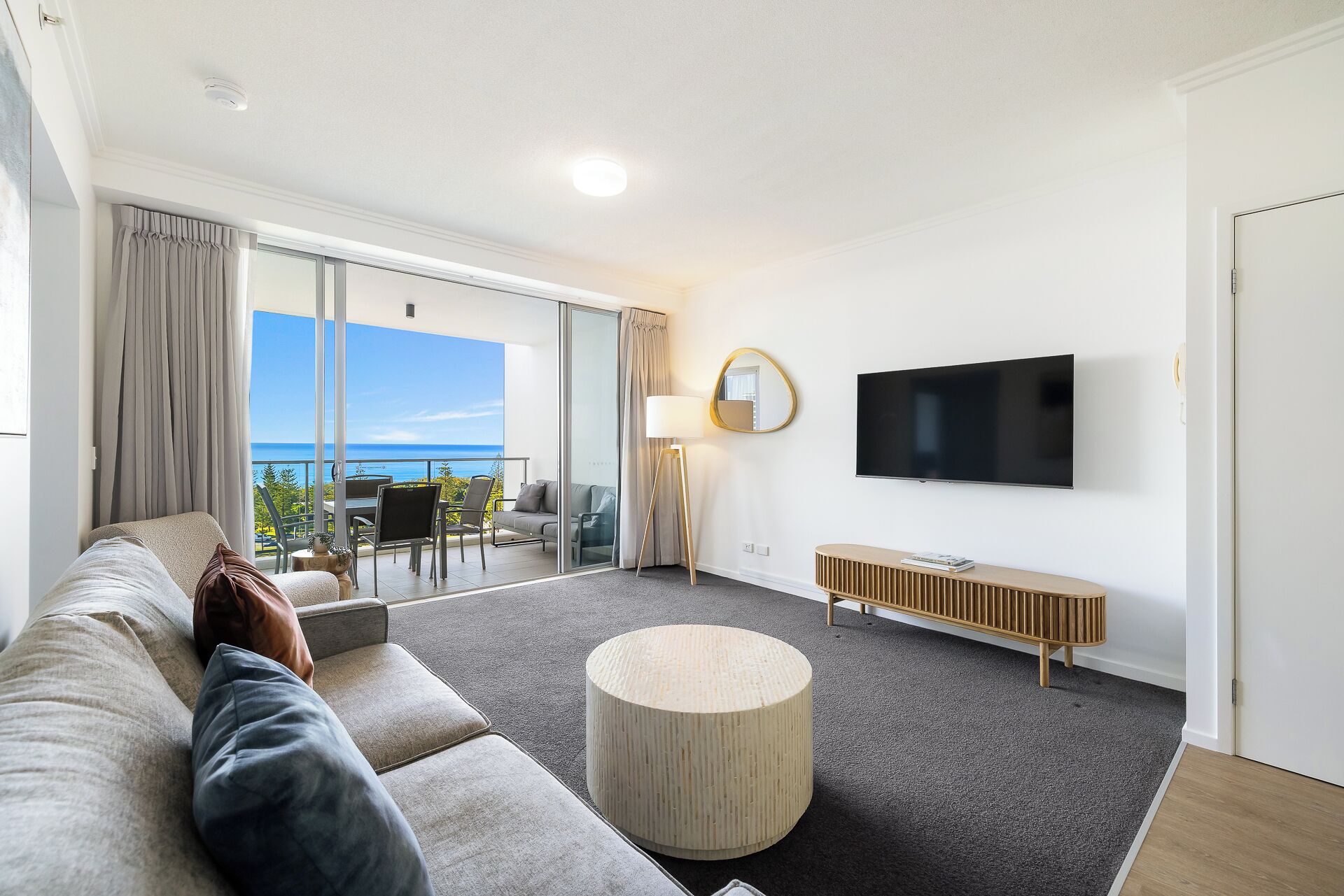 Een moderne woonkamer met een ruime bank, een ronde salontafel, een aan de muur bevestigde televisie en uitzicht op zee vanuit het balkon.