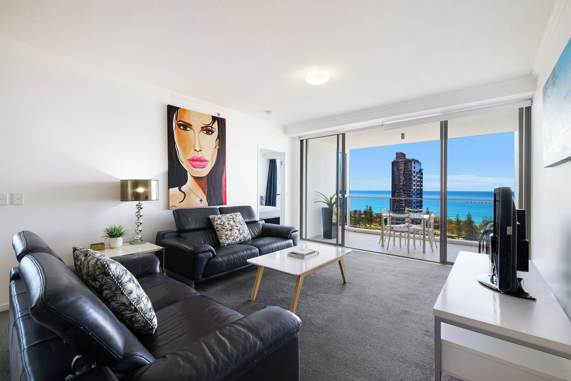 A modern living room with a black leather sofa, a coffee table, and a TV on a stand. There is a large window with a view of the ocean and a building.
