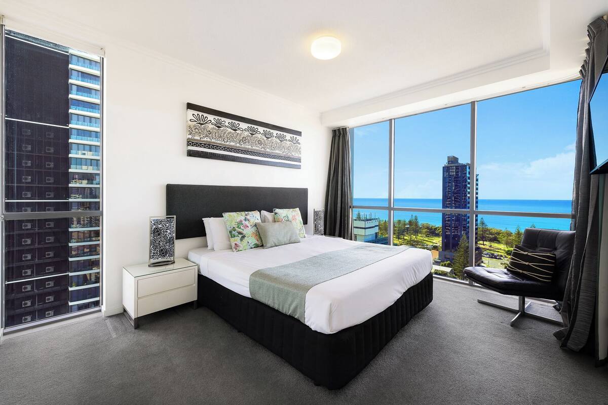 Una habitación de hotel moderna con una cama grande, una mesita de noche, un escritorio y una silla. La habitación tiene un ventanal amplio con vistas a la ciudad y al océano.