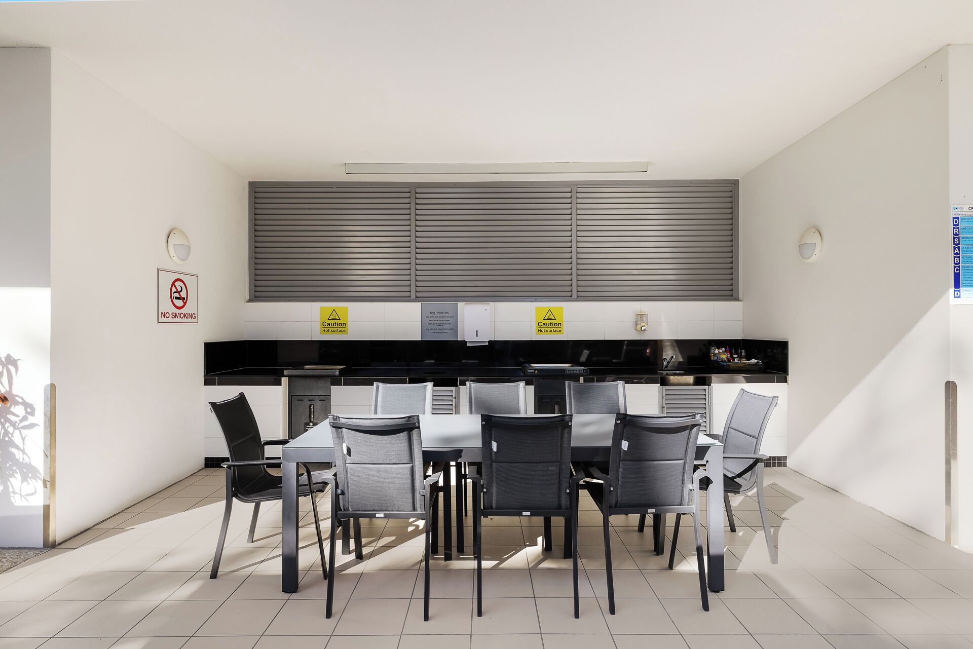 A modern kitchen with a dining area, featuring a 'No Smoking' sign, caution signs for hot surfaces, and a clean, minimalist design.