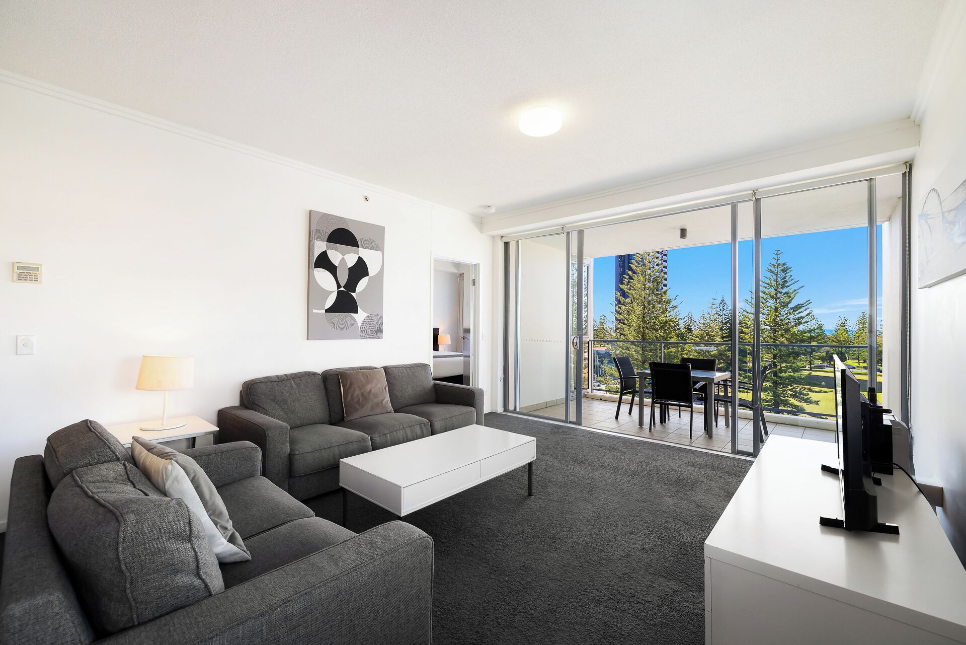 A modern living room with a grey sofa set, a white coffee table, and a TV on a white stand. There is a balcony with a glass door leading outside.