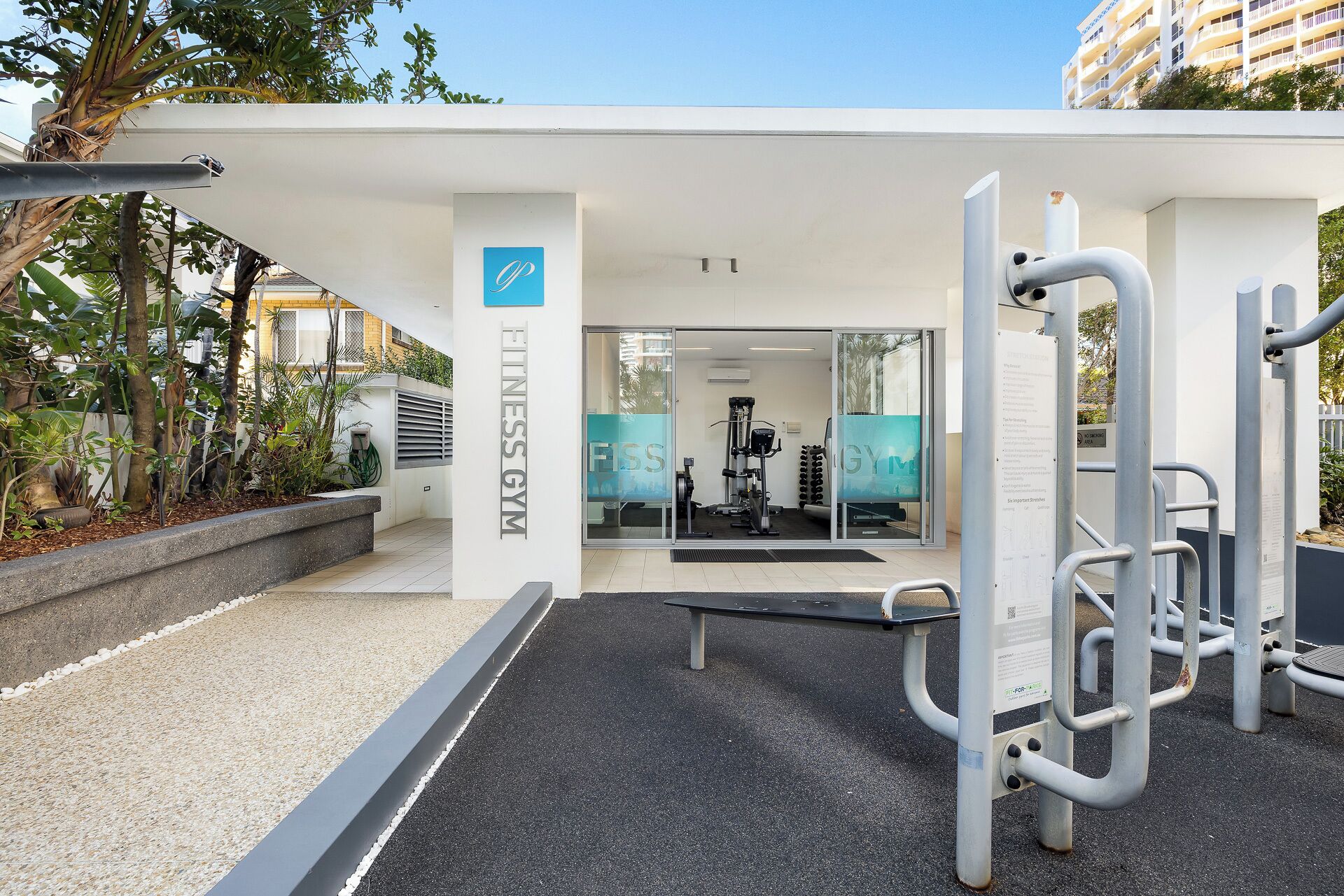 Zona de gimnasio al aire libre con equipo de ejercicio, un edificio blanco con letrero 'Fitness Gym' y un cielo despejado.