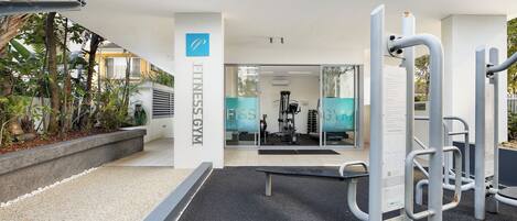 Zona de gimnasio al aire libre con equipo de ejercicio, un edificio blanco con letrero 'Fitness Gym' y un cielo despejado.